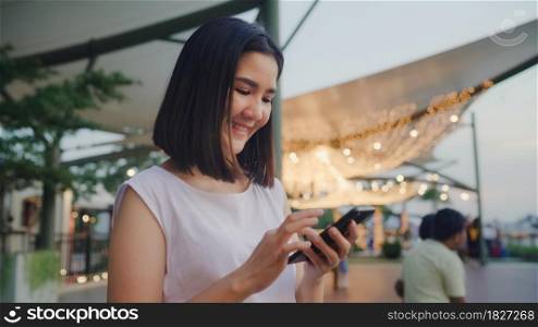 Happy young Asia woman blogger tourist with casual style hold smartphone slide screen on mobile phone with river view sunset near cafe in city town at night. Lifestyle tourist travel holiday concept.