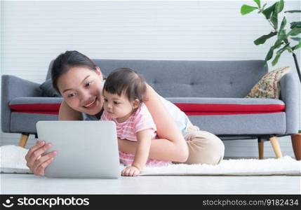 Happy young Asian beautiful mother sitting on floor, working with tablet while holding her cute little Caucasian 7 months newborn baby, taking care infant at home. Child care concept