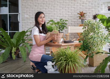 Happy young Asian beautiful woman wearing apron smiling and holding plant in wicker basket potted and take care of trees in greenhouse at home