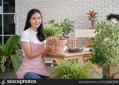 Happy young Asian beautiful woman wearing apron smiling and holding plant in wicker basket potted and take care of trees in greenhouse at home