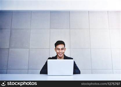 Happy Young Asian Businessman Working on Computer Laptop in his Workplace. Smiling and Looking at Computer Laptop. Sitting at the Desk in Industrial Loft Workplace. Wide Shot