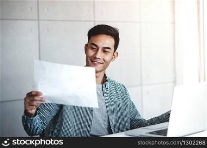 Happy Young Asian Businessman Working on Computer Laptop in his Workplace. Smiling and looking at Paper Worksheet