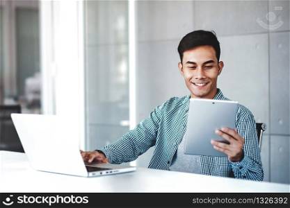 Happy Young Asian Businessman Working on Computer Laptop in his Workplace. Smiling and looking at Digital Tablet