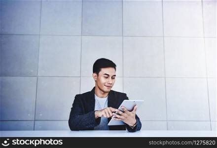 Happy Young Asian Businessman Working on Digital Tablet. Smiling and Sitting at the Desk in Industrial Loft Workplace. Wide Shot