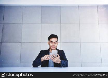 Happy Young Asian Businessman Working on Smartphone. Smiling and Sitting at the Desk in Industrial Loft Workplace. Wide Shot