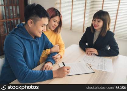 Happy young Asian couple and realtor agent. Cheerful young man signing some documents while sitting at desk together with his wife. Buying new house real estate. Signing good condition contract.