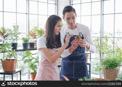 Happy young Asian couple in apron are planting plants in pots and looking after small cactus plant in shop together