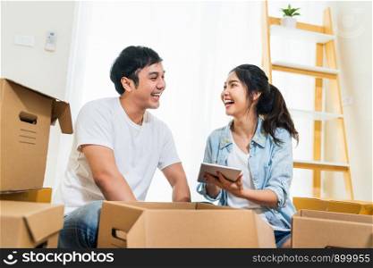 Happy young Asian couple moving in to new house, using digital tablet organizing things and unpacking boxes together. Home relocation, domestic lifestyle, or love relationship concept