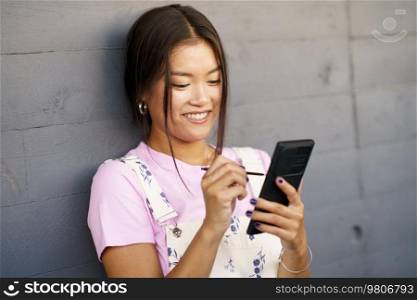 Happy young Asian female in stylish clothes smiling and using stylus to browse social media on cellphone while leaning on gray wall on city street. Cheerful Asian woman using smartphone near wall