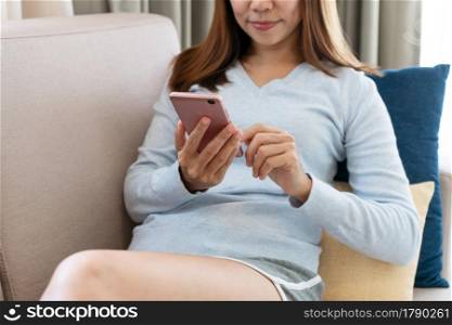 Happy young Asian female relaxing on couch at home and using smartphone. Closeup