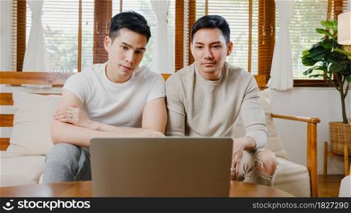 Happy young asian gay couple sit couch use laptop facetime video call with friends and family in living room at home. Stay at home quarantine, Social distancing, LGBTQ+ Young married concept.