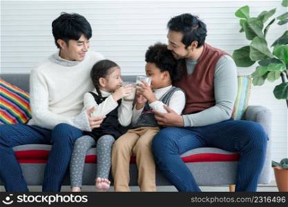 Happy young Asian gay couple with diverse adopted children African and Caucasian holding glass of milk sitting on sofa at home. Relationship of Lgbt family with kids son daughter foster concept.
