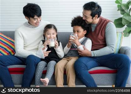 Happy young Asian gay couple with diverse adopted children, African and Caucasian, drinking milk and sitting on sofa at home. Relationship of Lgbt family with kids son daughter foster concept.