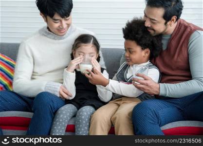 Happy young Asian gay couple with diverse adopted children, African and Caucasian, drinking milk and sitting on sofa at home. Older boy with milk mustache cheer up little girl drink milk. Lgbt family