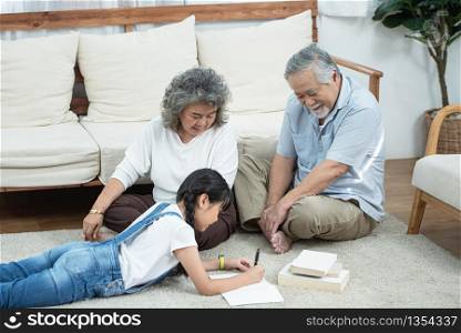 Happy young asian granddaughter reading and writing book with grandfather and grandmother looking beside on floor in living room at home,Retirement domestic life concept.
