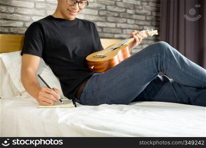 Happy young asian man playing ukulele sitting on bed in bedroom