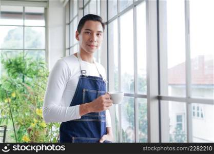 Happy young Asian man wearing apron smiling and holding cup of coffee looking at camera after take care of trees in greenhouse at home
