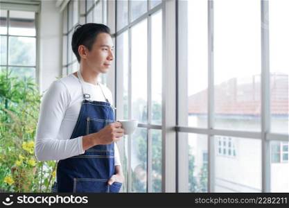 Happy young Asian man wearing apron smiling and holding cup of coffee looking out of window after take care of trees in greenhouse at home