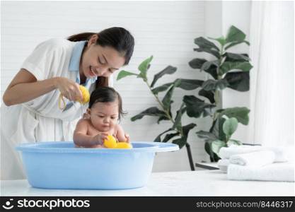 Happy young Asian mother bathing her little baby with sponge while cute daughter enjoy playing yellow duck toy and sitting in bathtub, Baby bathing at home concept. White background