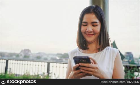 Happy young Asian woman blogger tourist with casual style hold smartphone look with smile joyful with beautiful sunset on river view near cafe in city town. Lifestyle tourist travel holiday concept.