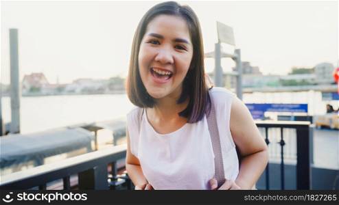 Happy young Asian woman blogger tourist with casual style looking at camera take selfie video call enjoy with beautiful sunset river view cafe in city town, Lifestyle tourist travel holiday concept.