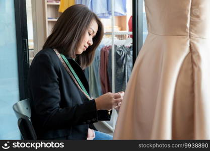 Happy young Asian woman dressmaker fashion designer is checking for completion for a suit and Dress in a showroom. Concept of success young entrepreneur in the fashion business.