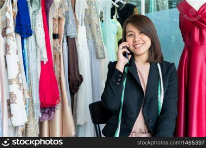 Happy young Asian woman dressmaker fashion designer uses a smartphone to accept orders from the customer in a showroom. Concept of success young entrepreneur in the fashion business.