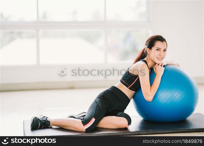Happy young Asian woman health exercise alone at home gym or sports club, rest on fitness ball. Yoga aerobic class, sport trainer, weight loss, or healthy wellbeing lifestyle concept. With copy space