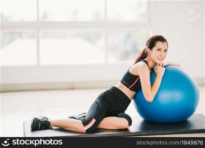 Happy young Asian woman health exercise alone at home gym or sports club, rest on fitness ball. Yoga aerobic class, sport trainer, weight loss, or healthy wellbeing lifestyle concept. With copy space