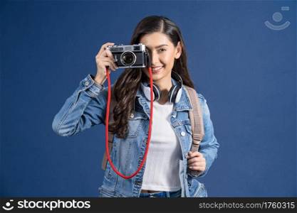 Happy young Asian woman holding and shooting photo with vintage camera on blue color background, carry backpacker and baggage, holiday and travel concept