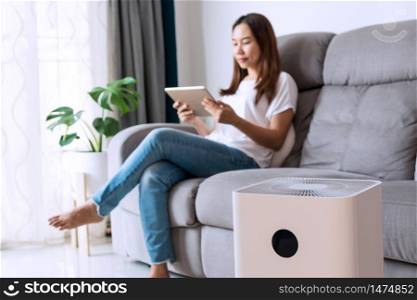 Happy young Asian woman relaxed on comfortable sofa at home, wellbeing breathing fresh air from air purifier while dust air pollution situation outside is really bad. PM 2.5, Healthy concept.