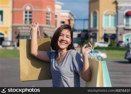 Happy young Asian woman shopping an outdoor market with background of pastel buildings and blue sky. Young asian woman smile with a colorful bag in her hand. Outdoor woman lifestyle shopping concept.
