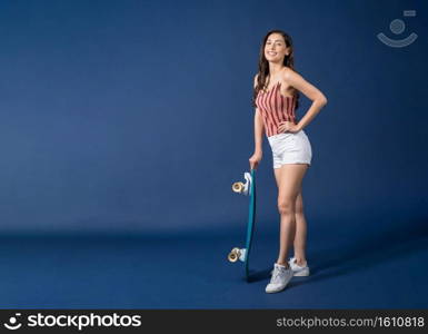 Happy young Asian woman standing and holding surfskate or skateboard on blue color background, exercise and sport, trendy and extream sports concept