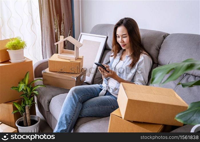 Happy young asian woman using smartphone in living room at new house with stack of cardboard boxes on moving day