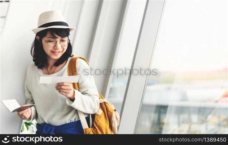 Happy Young Asian woman wear eyeglasses and hat holding her passport and ticket near the window of the airport.