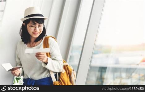 Happy Young Asian woman wear eyeglasses and hat holding her passport and ticket near the window of the airport.