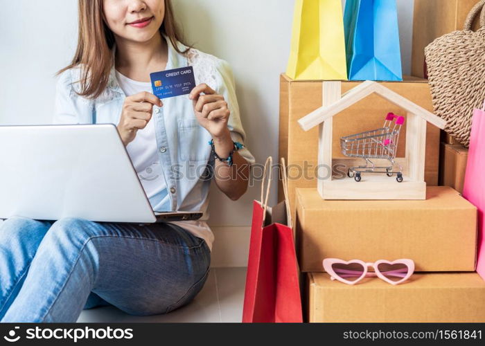 Happy young asian woman with colorful shopping bag, fashion items and stack of cardboard boxes at home, Using credit card for online shopping concept