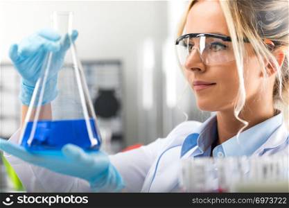Happy young attractive female scientist with protective eyeglasses and gloves holding a flask with blue liquid substance in the chemical research laboratory