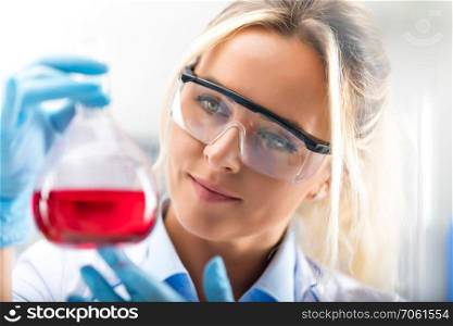 Happy young attractive female scientist with protective eyeglasses and gloves holding a flask with red liquid substance in the chemical research laboratory