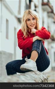 Happy young blond woman sitting on urban background. Smiling blonde girl with red shirt enjoying life outdoors.. Smiling blonde girl with red shirt enjoying life outdoors.