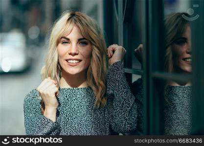 Happy young blond woman standing on urban background. Smiling blonde girl wearing dress outdoors.. Smiling young blonde woman standing on urban background.