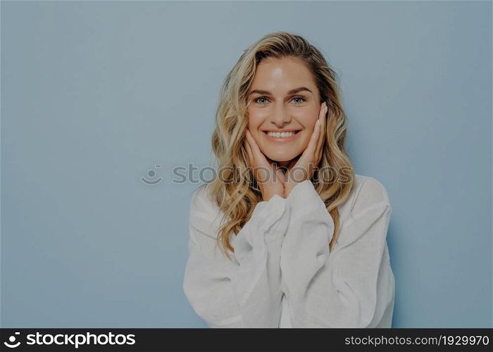 Happy young blonde female in casual outfit, gently touching face with both hands and smiling broadly, expressing happiness while standing isolated over blue studio background with copy space. Happy young blonde female gently touching face with both hands and smiling broadly