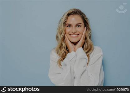 Happy young blonde female in casual outfit, gently touching face with both hands and smiling broadly, expressing happiness while standing isolated over blue studio background with copy space. Happy young blonde female gently touching face with both hands and smiling broadly