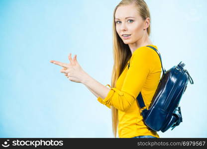 Happy young blonde teenage girl going to school or college wearing backpack pointing with finger at copy space. Studio shot on blue.. Young woman going to school