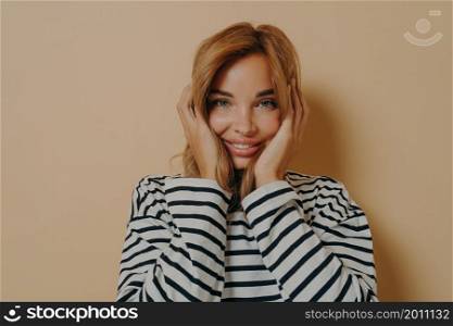 Happy young blonde woman looking at camera with joyful excited facial expression holding her face with both hands glad to hear good news, isolated over beige studio background. Happiness concept. Happy young woman looking at camera with joyful facial expression holding her face with both hands