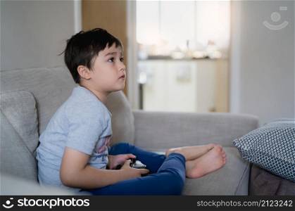 Happy young boy playing video games online with friends, Candid shot Cute child sitting on sofa holding game console.Portrait kid looking up at Monitor or TV while playing games and relaxing at home
