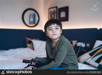 Happy young boy sitting in bed holding remote control, Cute Kid looking out with smiling face, Child relaxing at home watching TV in bed room.