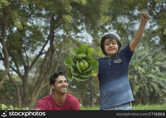 Happy young boy standing in garden holding a big cabbage in one hand and raising his fist with his father sitting beside him.