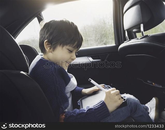 Happy young boy using a tablet computer while sitting in the back passenger seat of a car with a safety belt, Child boy drawing on smart pad