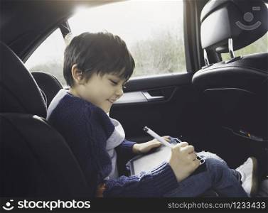 Happy young boy using a tablet computer while sitting in the back passenger seat of a car with a safety belt, Child boy drawing on smart pad